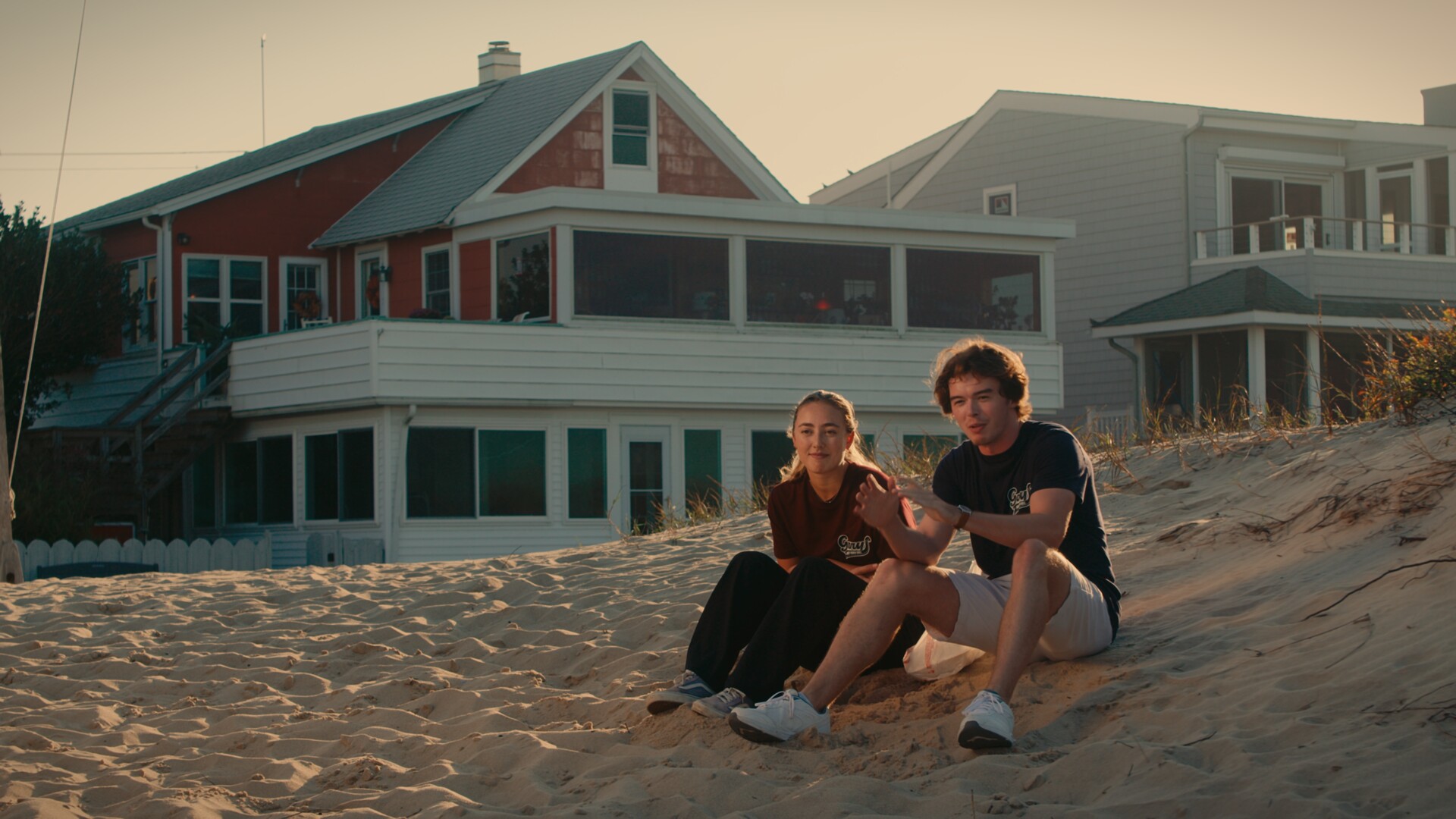 Two characters sitting on the dunes in front of a beach house at sunset